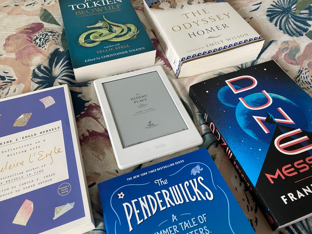 Books on a bedspread, including a Kindle version of The Hiding Place, The Odyssey, J. R. R. Tolkien's Beowulf, Dune Messiah, The Penderwicks