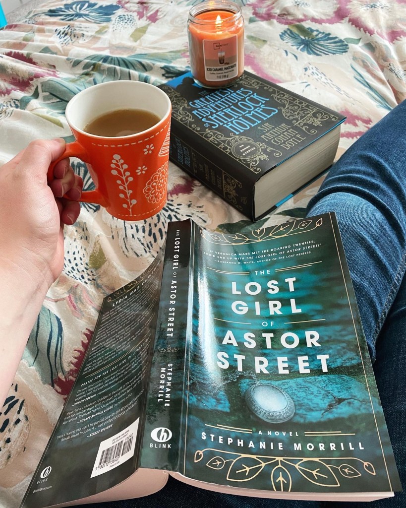 Cup of coffee, The Lost Girl of Astor Street book, and a collection of Sherlock Holmes, with a burning candle